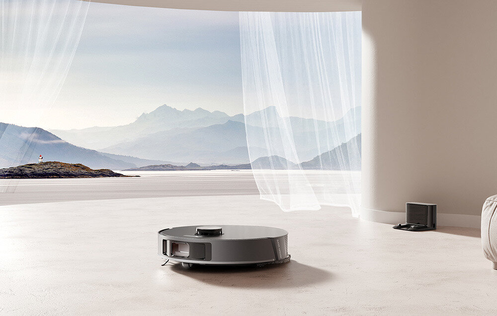 Na podłodze w jasnym pomieszczeniu znajduje się okrągły, szary robot sprzątający. W tle widać duże okno z widokiem na góry i jezioro, a obok stoi stacja dokująca.