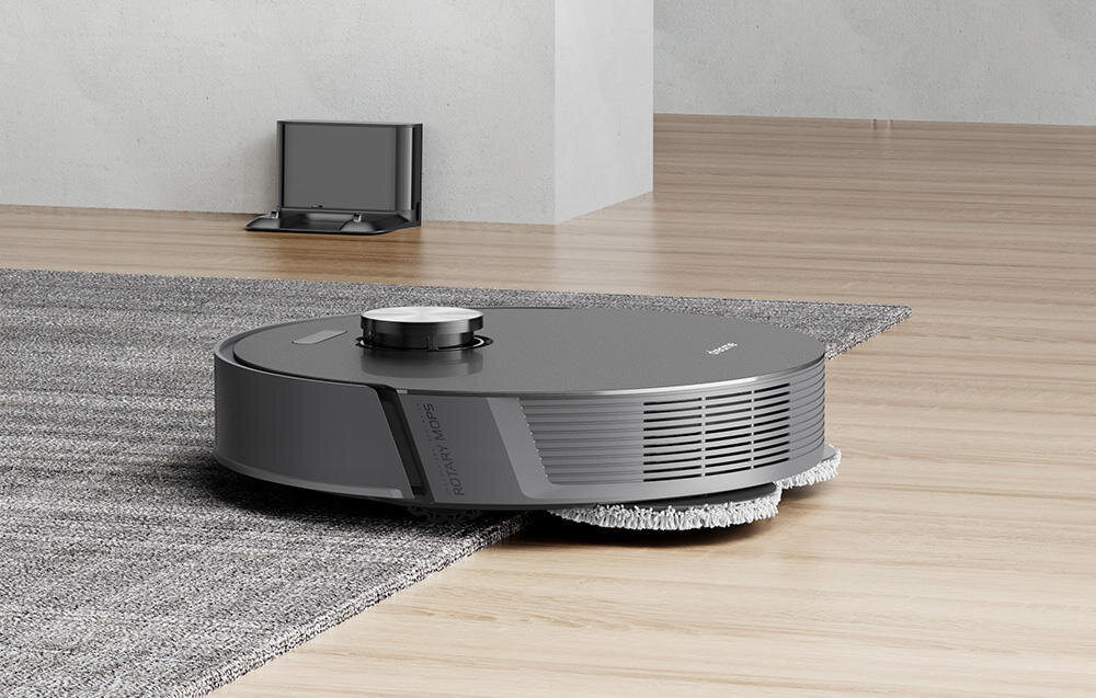 Na drewnianej podłodze znajduje się okrągły, ciemnoszary robot sprzątający z widocznymi szczotkami. W tle widać stację dokującą na jasnej podłodze.