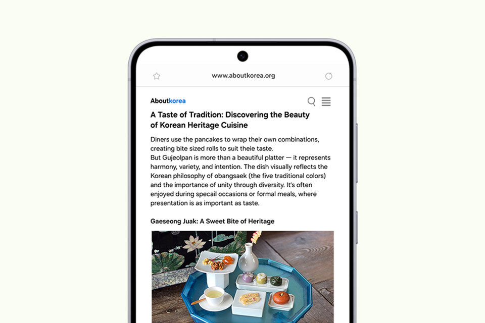 Na ekranie smartfona wyświetlany jest artykuł ze strony aboutkorea.org zatytułowany „Smak tradycji: odkrywanie piękna tradycyjnej kuchni koreańskiej”, w którym wyróżniono dania kuchni Gujeolpan i Gaseong Juak.