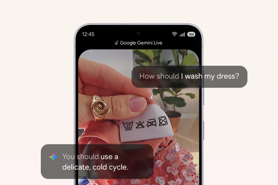 Ekran smartfona wyświetlający interfejs Google Gemini Live. Dłoń trzyma metkę z symbolami prania. Na ekranie pojawia się pytanie: „Jak prać sukienkę?” i odpowiedź: „Należy używać delikatnego programu do prania w zimnej wodzie”. W tle widoczna jest sukienka z cekinami i roślina.