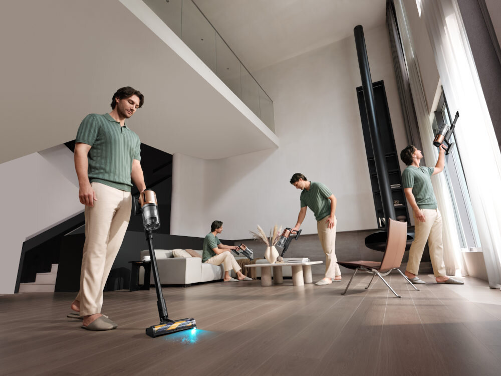 Wnętrze nowoczesnego salonu z dużymi oknami i drewnianą podłogą. Kilka osób używa odkurzacza pionowego na podłodze i przy oknie.