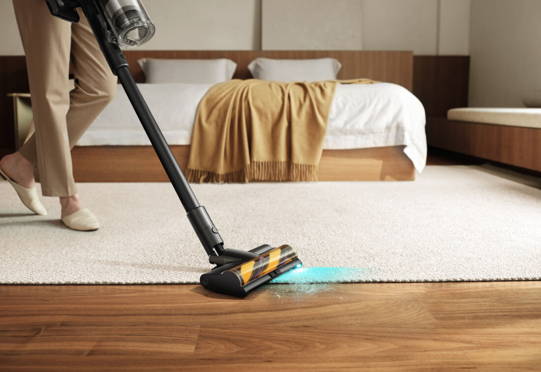 Odkurzacz na drewnianej podłodze i dywanie w sypialni. W tle łóżko z pościelą i kocem.