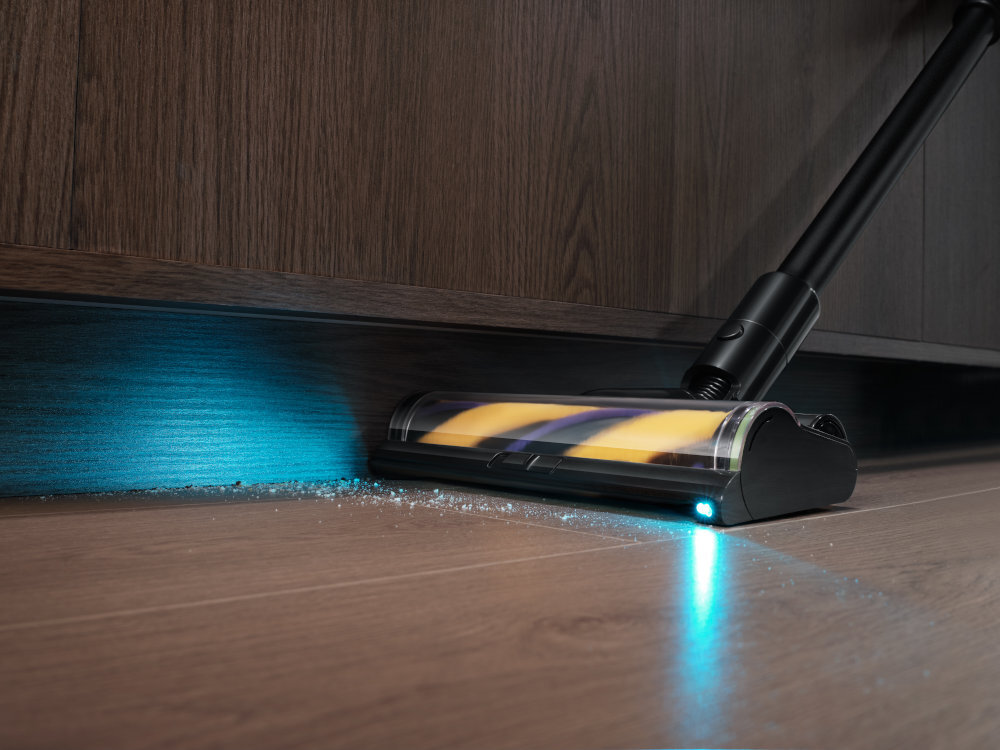 Odkurzacz z podświetleniem LED zbiera drobinki z podłogi przy drewnianej szafce. Widoczna jest szczotka z kolorowymi elementami wewnątrz przezroczystej obudowy.