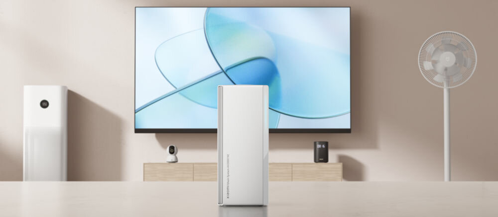 Router XIAOMI Mesh System AX3000 NE 2.4 / 5 GHz (DualBand), Wi-Fi Mesh (1 szt.) prezentacja routera na stole w tle na beżowej ścianie powieszony telewizor pod nim regał z urządzeniami obok klimator i wiatrak szerokie pasmo 160 MHz wysoka przepustowość