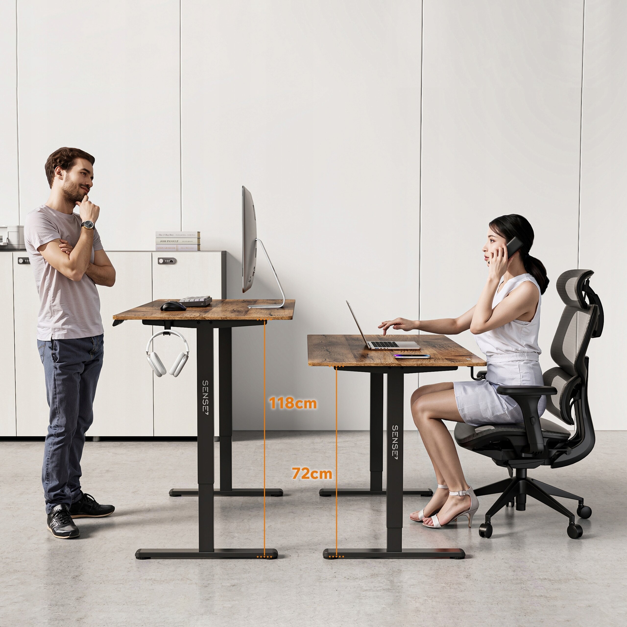 Dwa biurka z ciemnobrązowym blatem i czarnymi nogami, jedno ustawione na wysokości 118 cm, drugie na 72 cm. Na biurkach znajdują się komputer i laptop, a na nogach widoczne jest logo \'SENSE7\'.