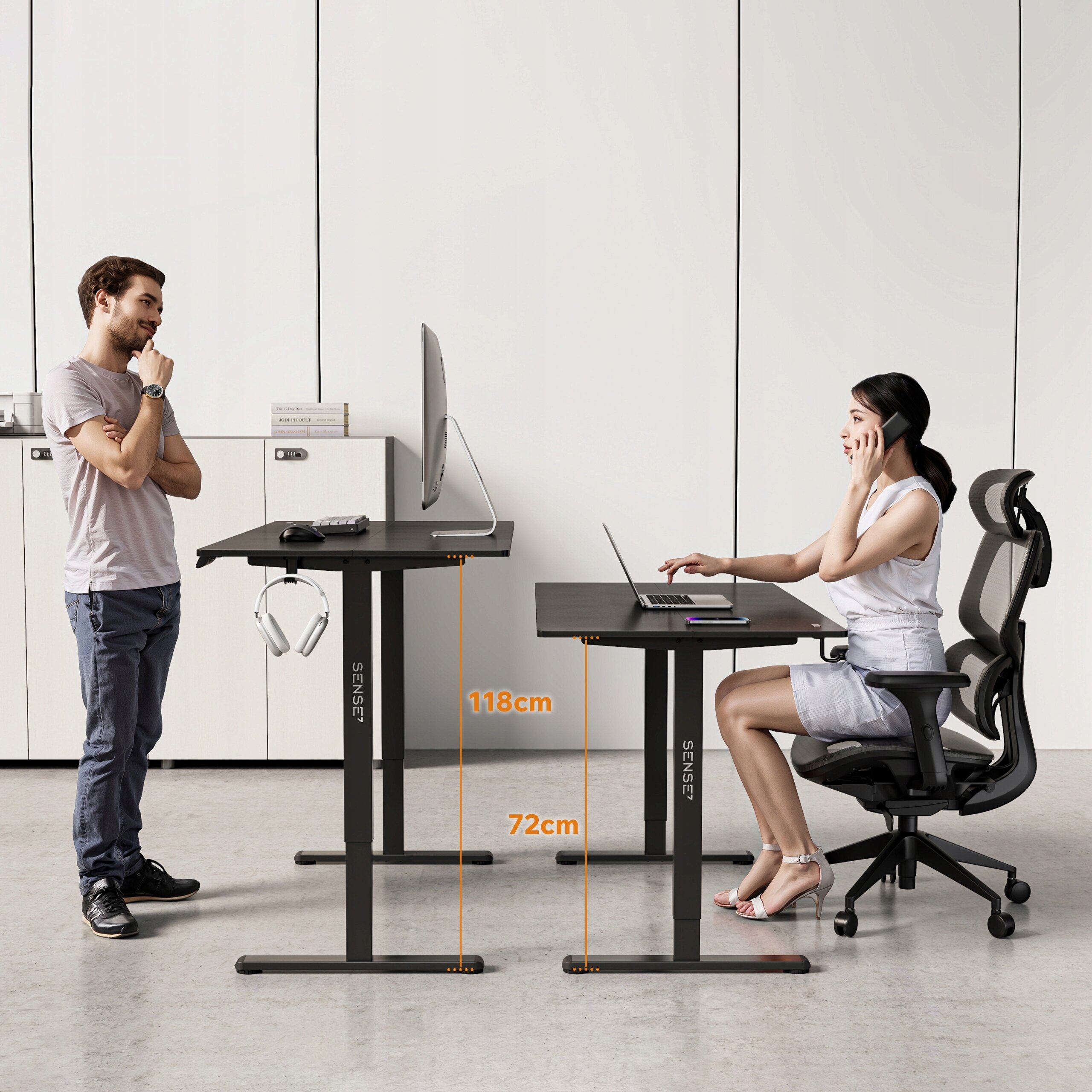 Na obrazie widoczne są dwa biurka komputerowe z regulacją wysokości, jedno ustawione na 118 cm, a drugie na 72 cm. Na biurkach znajdują się monitory i akcesoria komputerowe, a na jednym z nich wisi para słuchawek; na nogach biurek widnieje napis \'SENSE7\'.