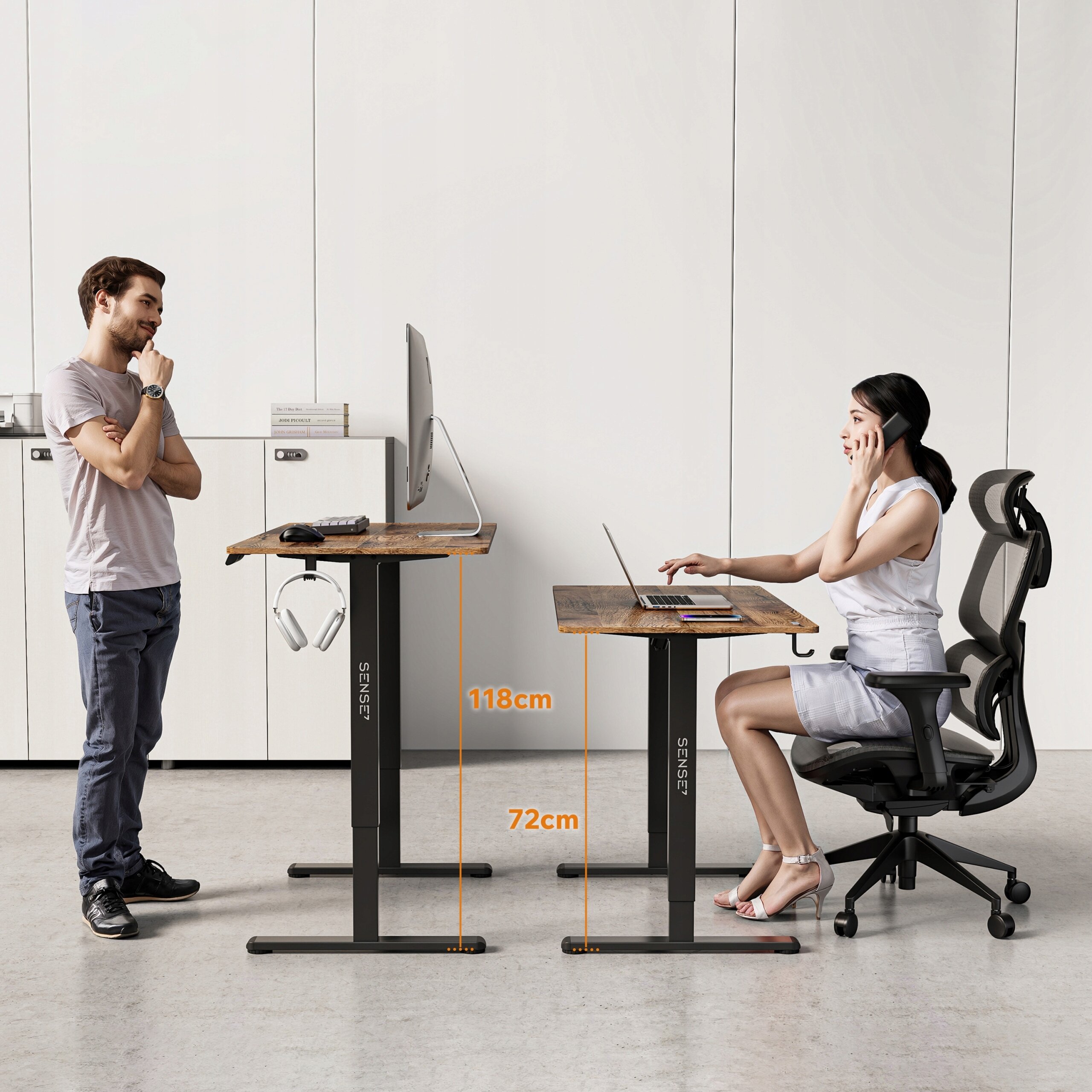 Na obrazie widoczne są dwa biurka regulowane elektrycznie z oznaczeniami wysokości 118 cm i 72 cm. Na biurkach znajdują się monitory, a przy jednym z nich siedzi osoba na krześle biurowym, rozmawiająca przez telefon.