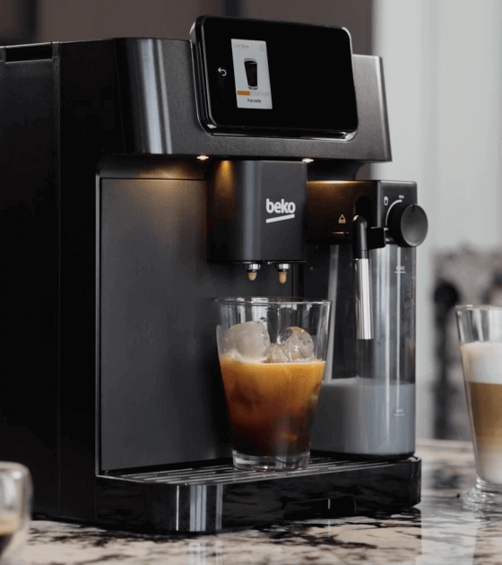 Czarny ekspres do kawy z logo \'beko\' przygotowuje kawę mrożoną w szklance z lodem. Obok znajduje się pojemnik na mleko oraz szklanka z kawą z mlekiem.