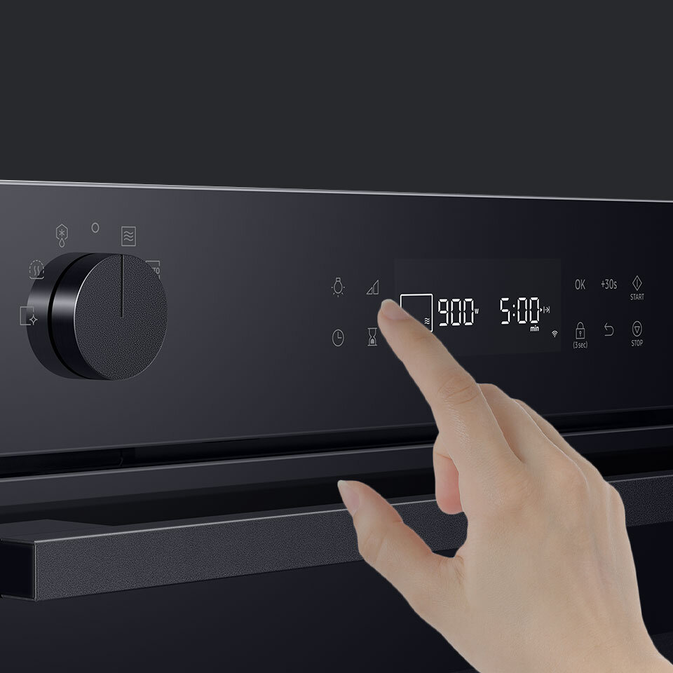 Na obrazie widoczny jest panel sterowania kuchenki mikrofalowej z wyświetlaczem pokazującym \'900W\' i \'5:00 min\'. Dłoń dotyka ekranu, a obok znajduje się pokrętło oraz kilka ikon funkcyjnych.