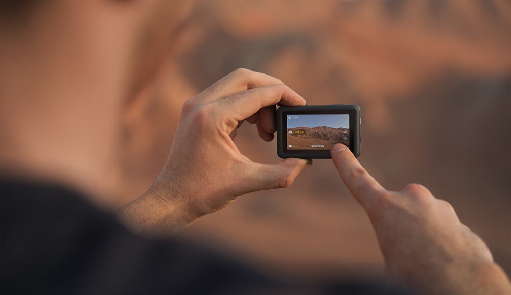 Kamera sportowa DJI Osmo Action 6 Dłonie trzymają małą kamerę sportową kadrującą skalisty krajobraz na wyświetlaczu, aplikacja, narzędzia, podgląd