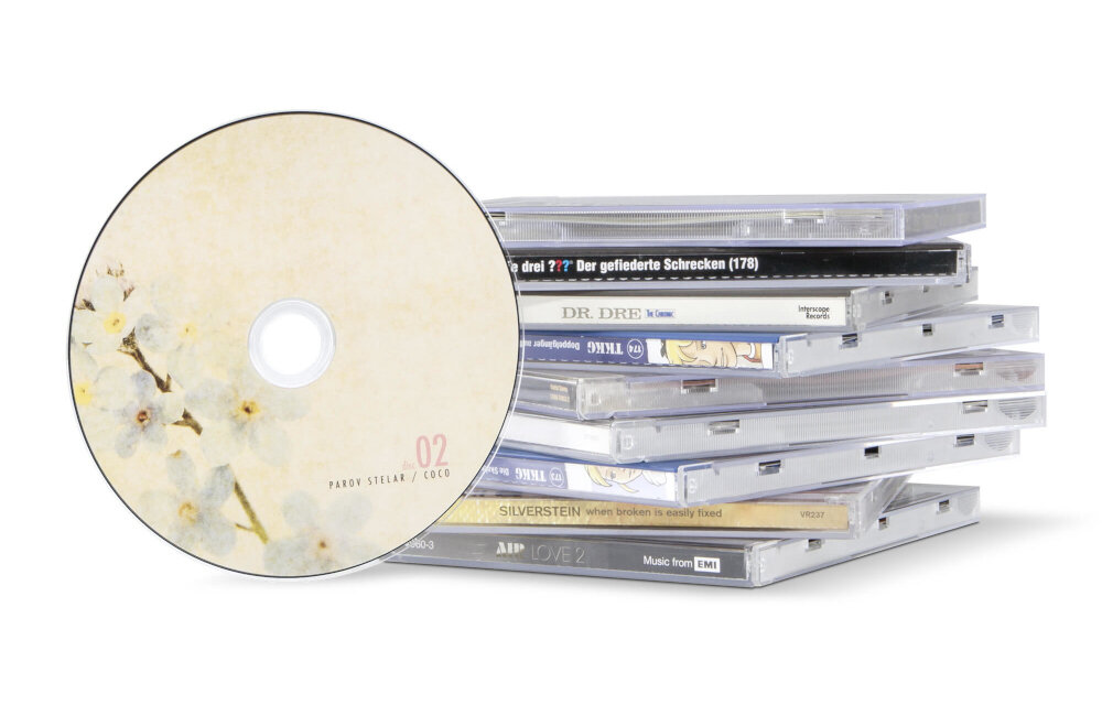 Na obrazie widoczna jest płyta CD z kwiatowym wzorem i napisem \'02 PAROV STELAR / COCO\', obok stosu pudełek na płyty CD z widocznymi tytułami, takimi jak \'DR. DRE\' i \'SILVERSTEIN\'.