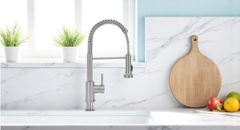 Na obrazie widoczna jest nowoczesna bateria kuchenna w kolorze niklu szczotkowanego, zamontowana na marmurowym blacie. W tle znajdują się dwa doniczki z roślinami, okno oraz drewniana deska do krojenia i owoce.