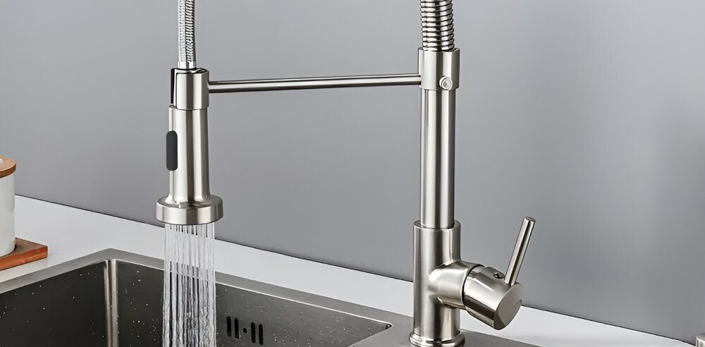 Na obrazie widoczna jest metalowa bateria kuchenna z elastycznym wężem i uchwytem, zamontowana nad zlewem. Woda wypływa z wylewki, a powierzchnia baterii ma wykończenie w kolorze niklu szczotkowanego.