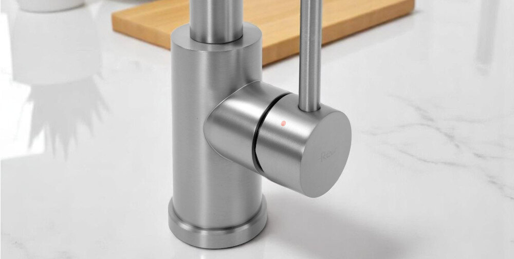 Na obrazie widoczna jest metalowa bateria w kolorze niklu szczotkowanego, zamontowana na blacie z marmurowym wzorem. Na uchwycie baterii znajduje się mały czerwony punkt oraz napis \'Rea\'.