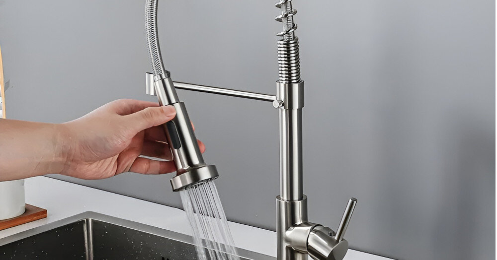 Osoba trzyma w ręku słuchawkę prysznicową z baterii kuchennej, z której wypływa woda. Bateria wykonana jest z niklu szczotkowanego i ma nowoczesny design.