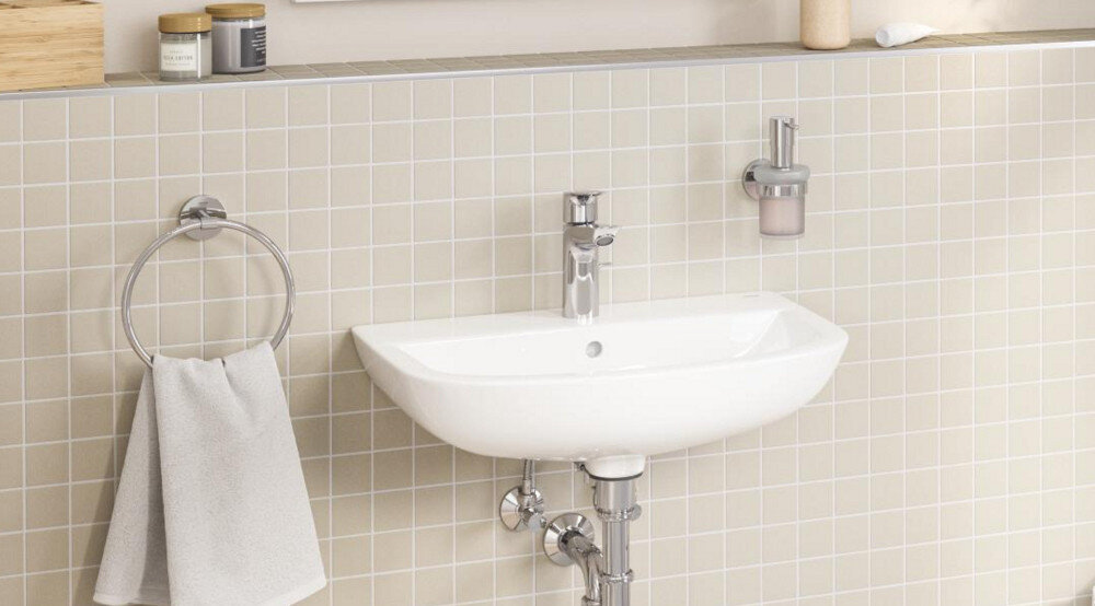 DOZOWNIK GROHE ESSENTIALS 40394001 CHROM Biała umywalka na tle jasnych płytek z chromowaną baterią, obok uchwyt na ręcznik i dozownik mydła – minimalistyczne wyposażenie łazienki