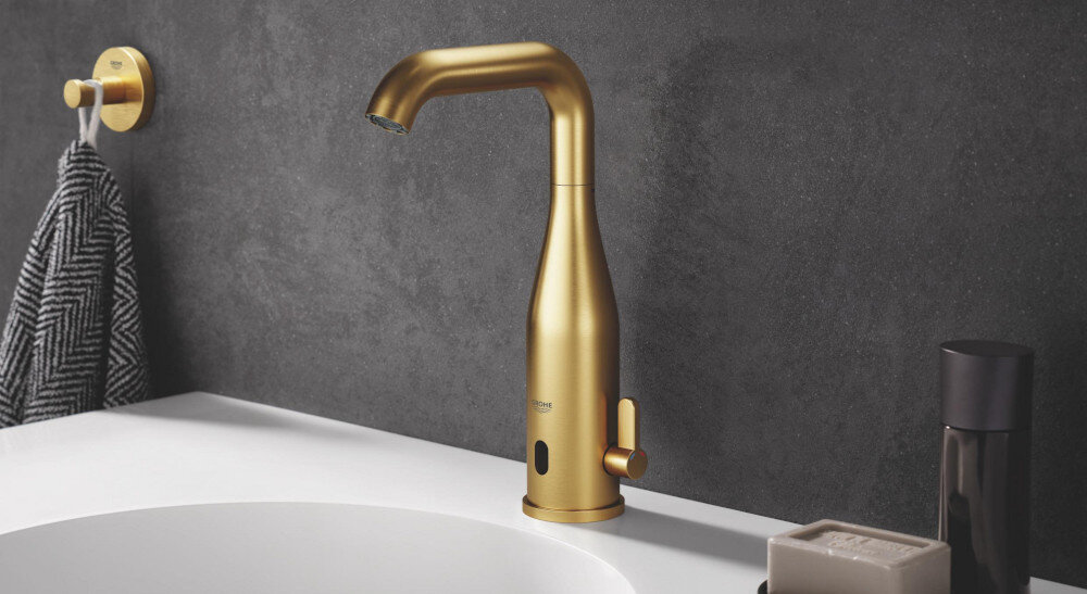 HACZYK NA PŁASZCZ KĄPIELOWY GROHE ESSENTIALS 40364GN1 Miedź szczotkowana Na umywalce w łazience zamontowana jest złota bateria z czujnikiem, ustawiona na tle ciemnej, teksturowanej ściany. Obok znajdują się akcesoria łazienkowe, takie jak dozownik i mydelniczka, a na ścianie wisi uchwyt z ręcznikiem w podobnej kolorystyce
