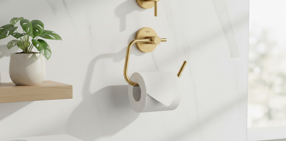 UCHWYT NA PAPIER GROHE ESSENTIALS 40689GN1 Złoty uchwyt na papier toaletowy zamontowany na jasnej, marmurowej ścianie; obok widoczna półka z rośliną, całość w minimalistycznym stylu