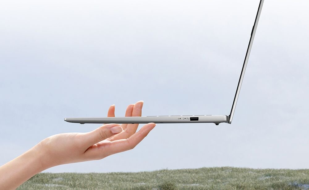 Osoba trzyma cienki, srebrny laptop w powietrzu, na tle nieba i trawy. Laptop jest otwarty, a jego profil jest smukły.