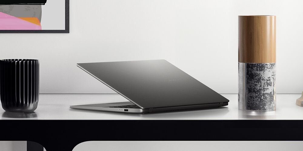 Na biurku znajduje się otwarty laptop w kolorze ciemnym, obok czarny kubek i dekoracyjny pojemnik z drewnianą pokrywką. W tle widoczny jest fragment obrazu na ścianie.