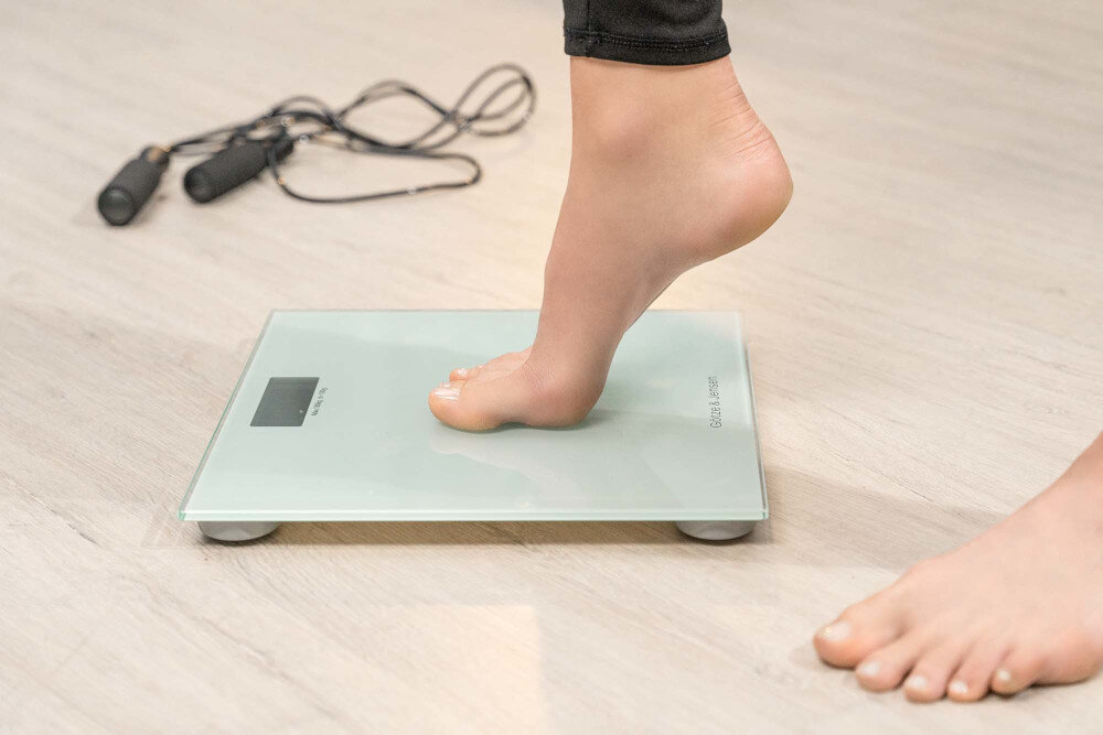 Osoba stoi na szklanej wadze łazienkowej z widocznym logo \'Götze & Jensen\'. W tle leży skakanka na drewnianej podłodze.