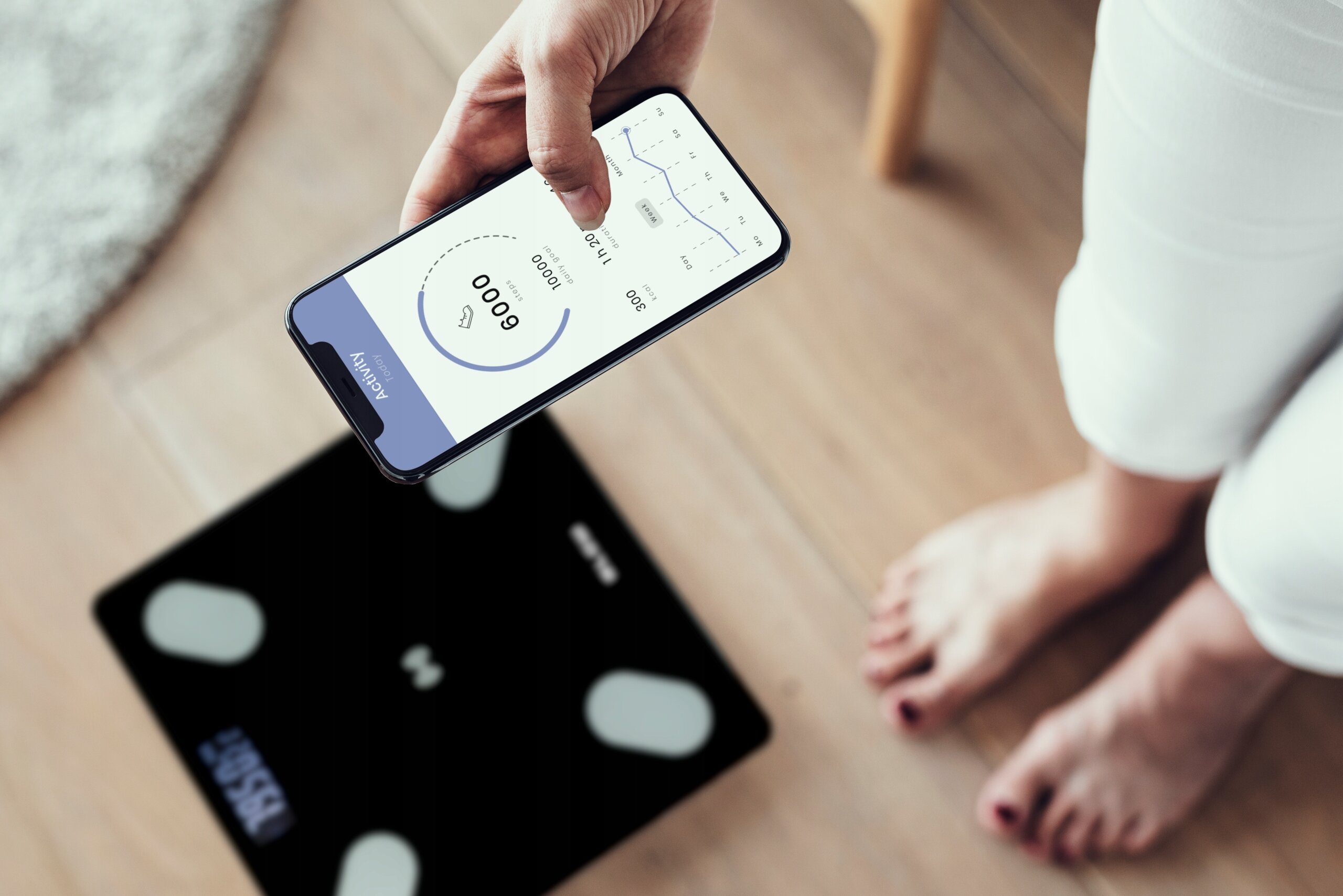 Osoba trzyma smartfon z aplikacją pokazującą dane analityczne, a na podłodze widoczna jest czarna waga łazienkowa z czterema srebrnymi sensorami.