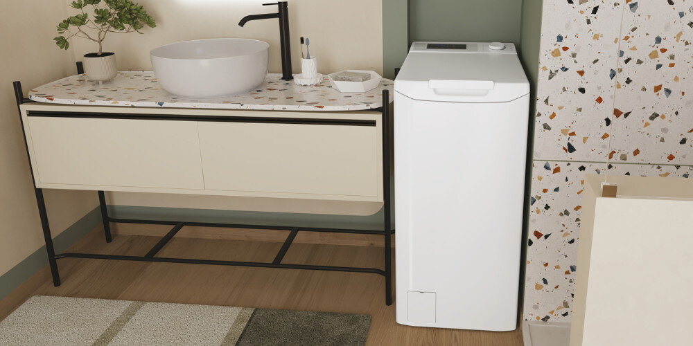 Wnętrze łazienki z białą pralką ładowaną od góry obok umywalki na blacie z kolorowym wzorem. Ściany i podłoga są w neutralnych kolorach, a na blacie znajdują się akcesoria łazienkowe.