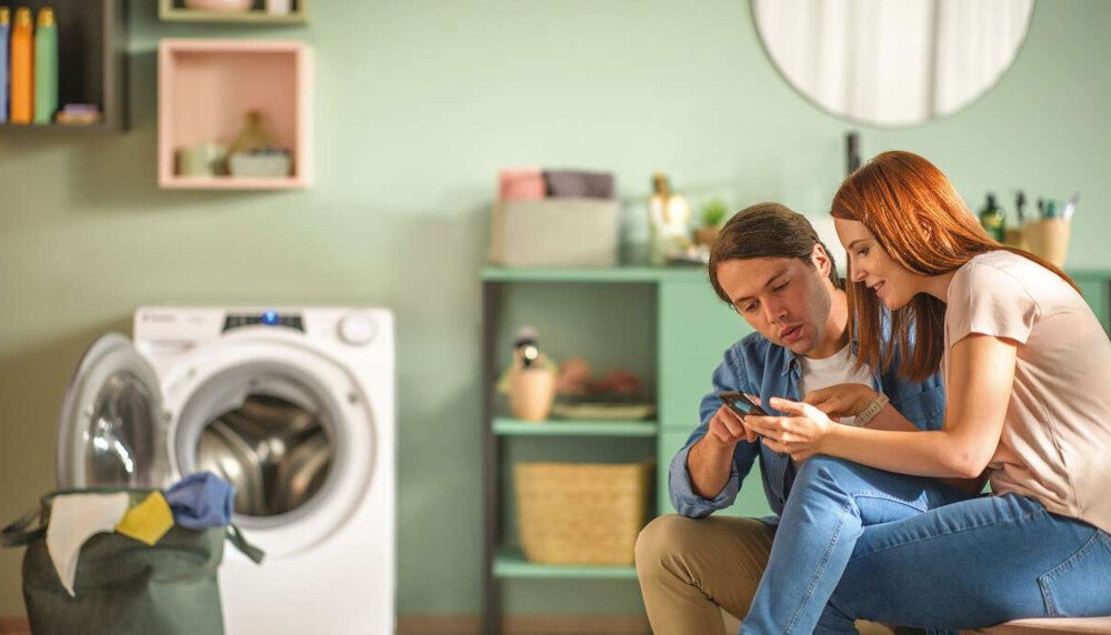 W pomieszczeniu znajduje się pralka z otwartymi drzwiczkami, obok której stoi kosz z praniem. W tle widoczna jest półka z różnymi przedmiotami.