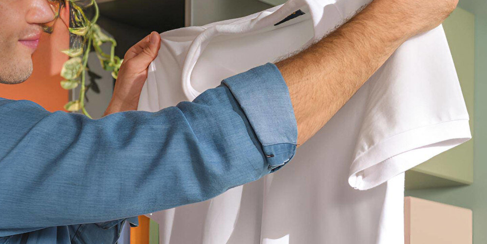 Osoba trzyma białą koszulkę w jasnym pomieszczeniu, w tle widoczne są rośliny i kolorowe ściany.