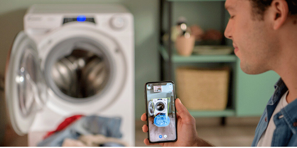 Osoba trzyma smartfon z wyświetlonym zdjęciem pralki, w tle widoczna jest otwarta pralka z ubraniami. W pomieszczeniu znajduje się zielona szafka z koszem i dekoracjami.