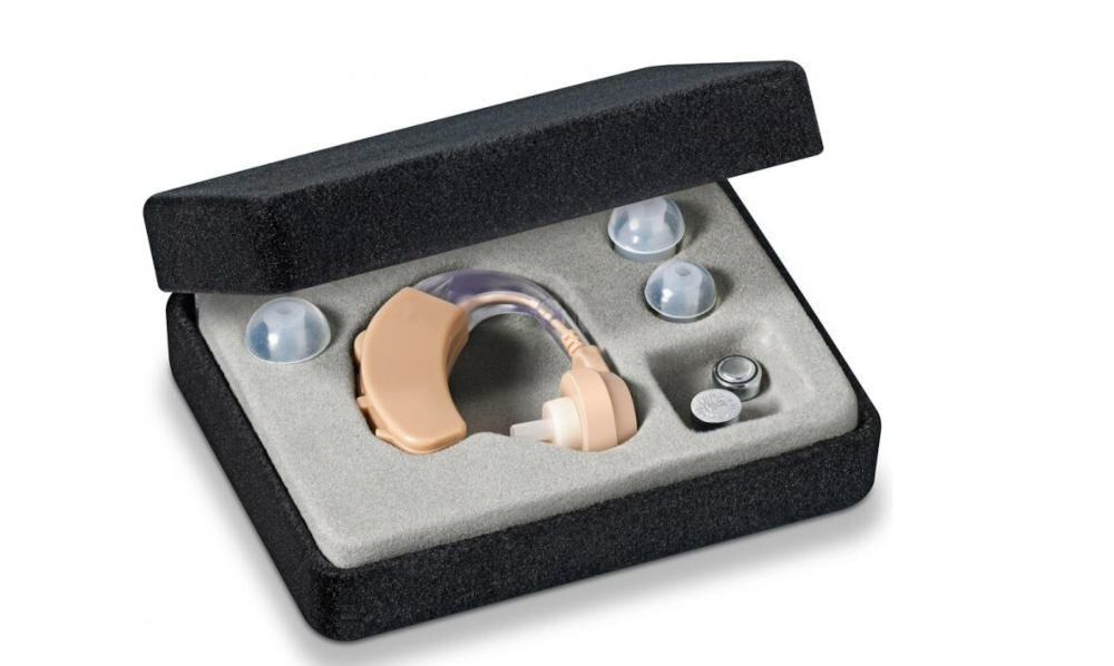Na obrazie widoczny jest aparat słuchowy w otwartym etui, wraz z trzema końcówkami i dwoma bateriami. Etui jest wyłożone jasnym materiałem, a aparat ma beżowy kolor.