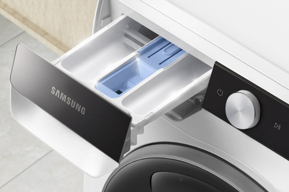 Zdjęcie przedstawia otwarty dozownik na detergenty w pralce z widocznym logo \'SAMSUNG\'. Obok znajduje się panel sterowania z pokrętłem i przyciskiem z symbolem włączania.