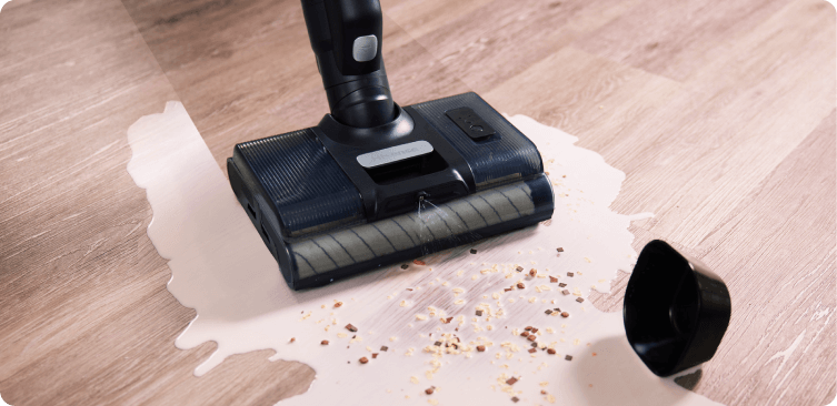 Odkurzacz czyszczący rozlany płyn i rozsypane drobinki na drewnianej podłodze. Obok leży czarna nakładka.
