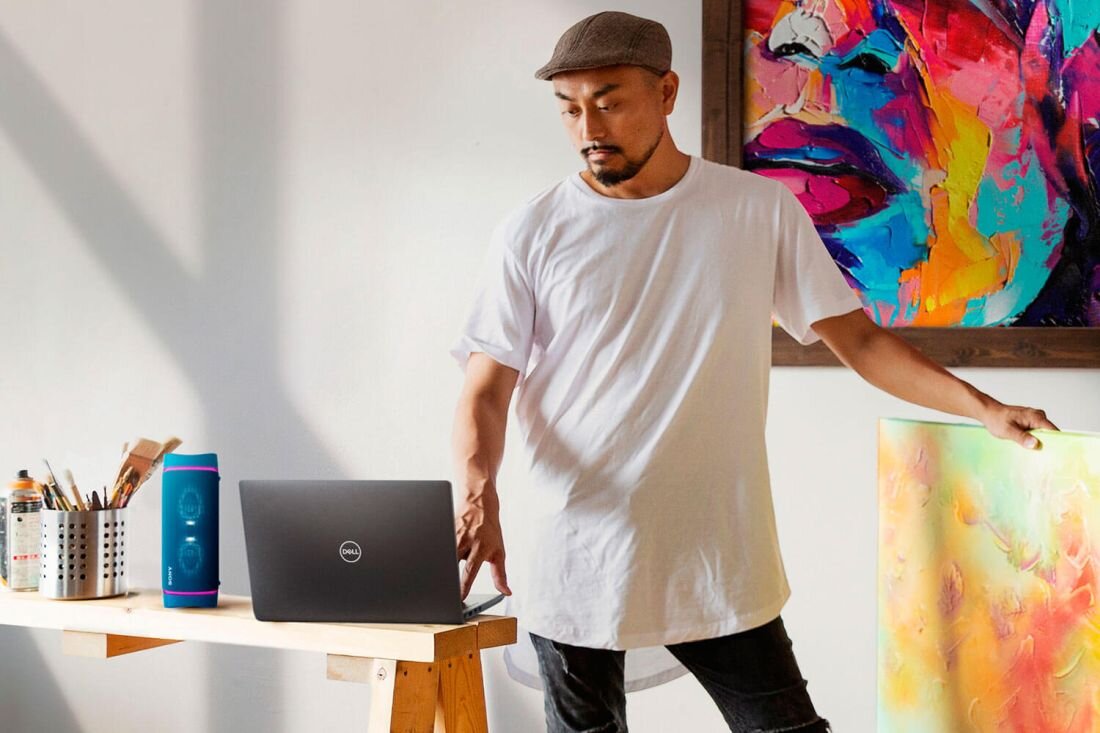 Na drewnianym stole stoi otwarty laptop z logo DELL, obok którego znajduje się niebieski głośnik. W tle widać kolorowy obraz oraz osobę w białej koszulce i czapce.