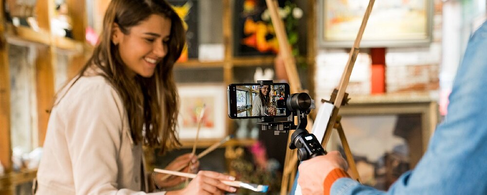 Osoba maluje na płótnie, podczas gdy inna osoba trzyma gimbal z zamocowanym smartfonem, który rejestruje scenę. W tle widoczne są obrazy i elementy dekoracyjne.