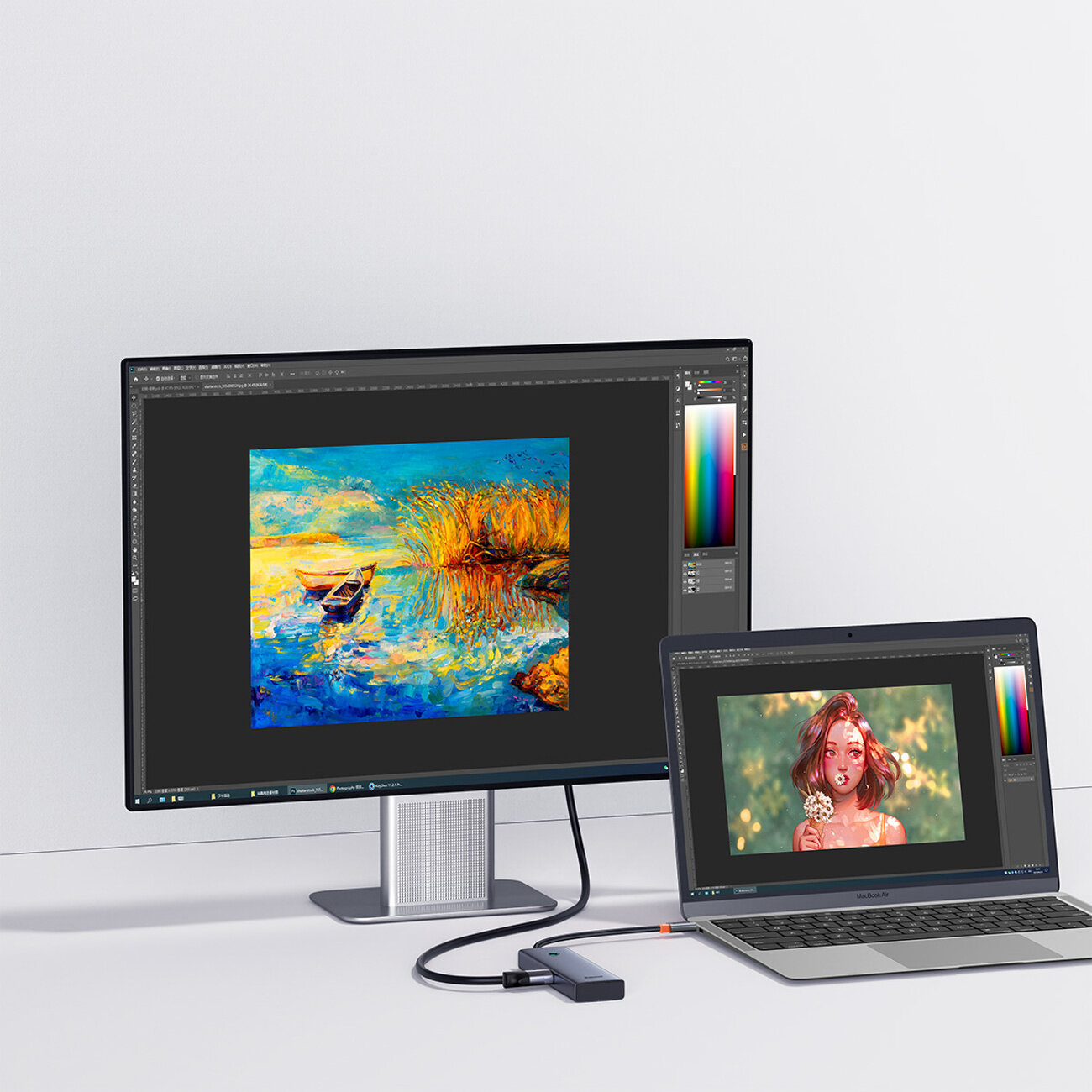 Na biurku znajduje się monitor i laptop, oba wyświetlają różne obrazy graficzne. Do laptopa podłączony jest kompaktowy hub USB-C z kilkoma portami USB-A i HDMI.