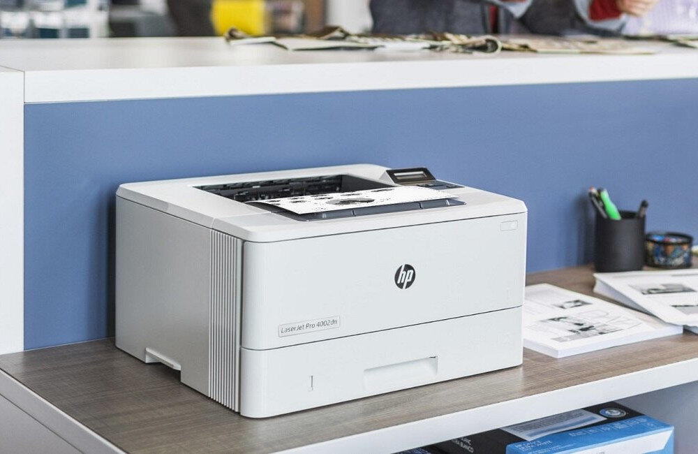 Na biurku stoi drukarka z logo HP, model LaserJet Pro 4002dn. Obok drukarki znajdują się dokumenty i przybory biurowe.