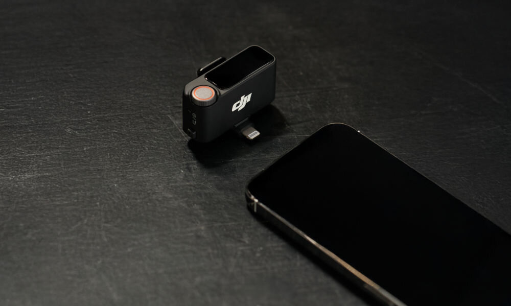 Na ciemnym tle znajduje się adapter z logo DJI oraz smartfon. Adapter ma złącze Lightning i okrągły przycisk na górze.
