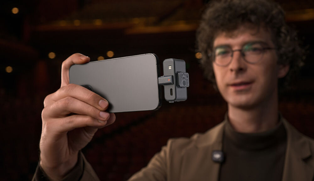 Osoba trzyma smartfon z podłączonym adapterem DJI Mic 3 Lightning. W tle widoczne jest rozmyte wnętrze budynku.