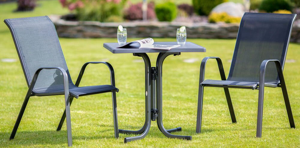 Na trawniku stoją dwa czarne krzesła ogrodowe z metalowymi ramami i siatkowym oparciem, obok kwadratowego stolika z dwoma szklankami z napojem i otwartą gazetą. W tle widoczny jest kamienny mur i roślinność.