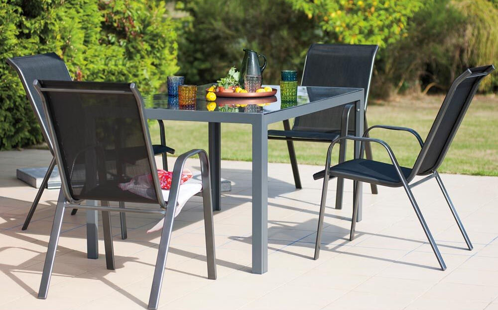 Na obrazie widoczny jest zestaw mebli ogrodowych składający się z czterech czarnych krzeseł i stołu z przezroczystym blatem. Na stole znajdują się różnokolorowe szklanki, dzbanek oraz owoce.