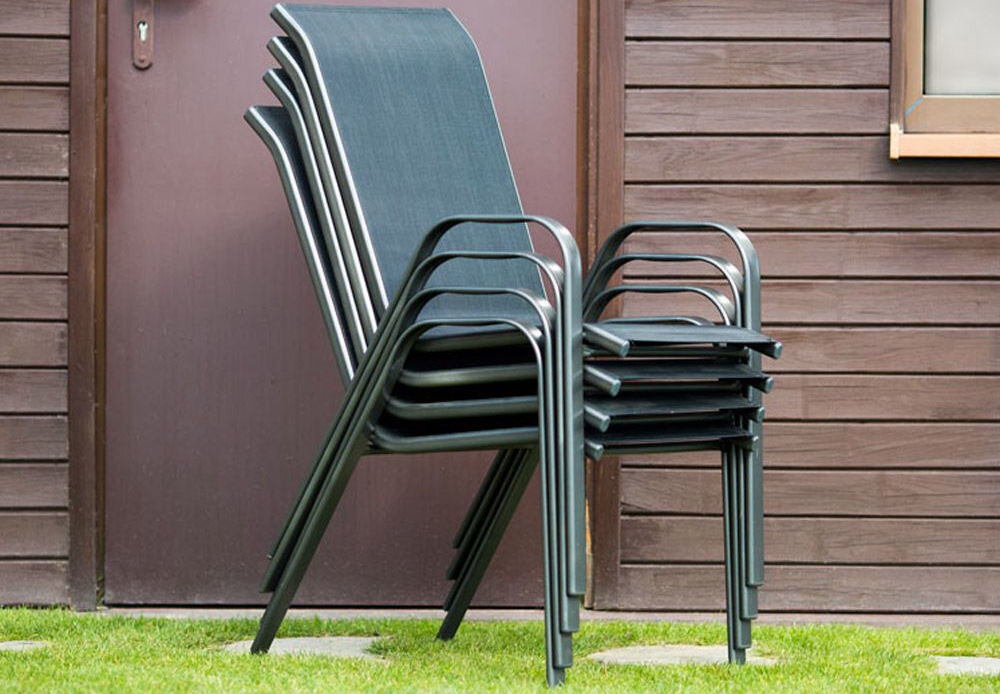 Na obrazie widoczne są czarne krzesła ogrodowe ustawione jedno na drugim przed drewnianą ścianą budynku. Krzesła mają metalowe ramy i ciemne siedziska.