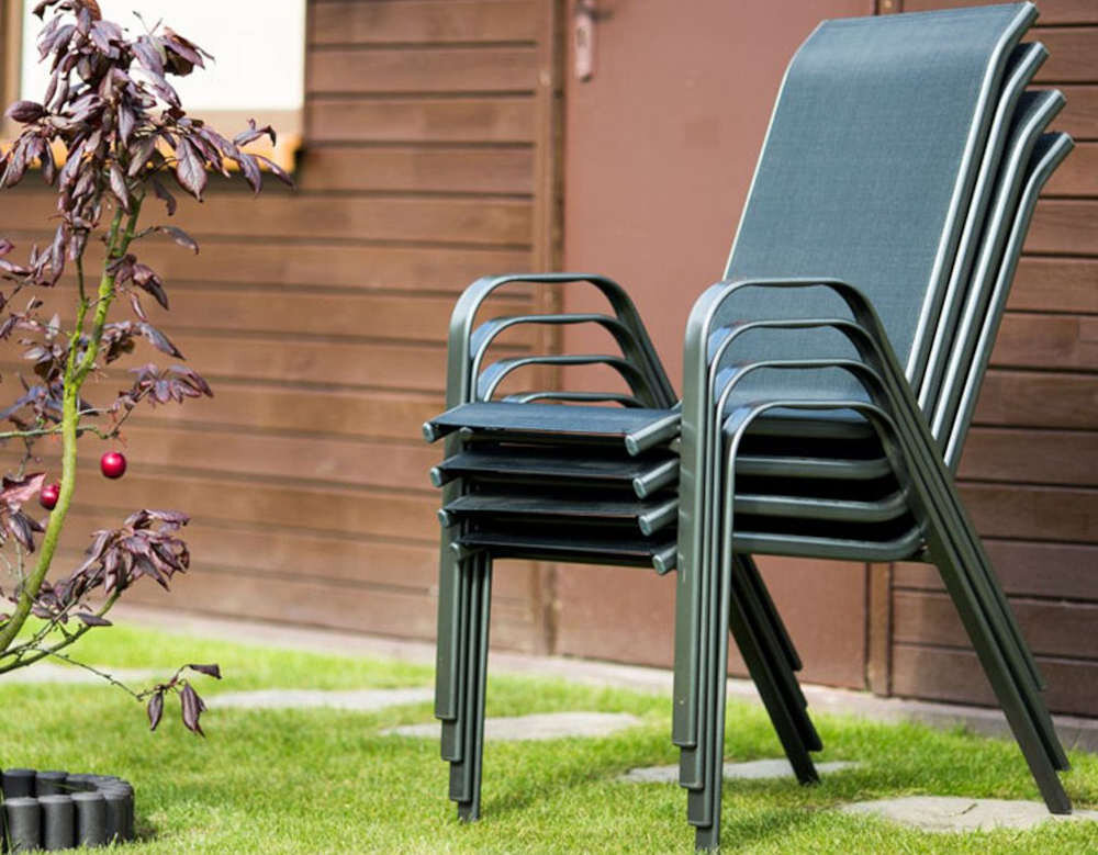 Na trawniku stoi stos kilku czarnych krzeseł ogrodowych z metalowymi ramami. W tle widoczna jest drewniana ściana budynku oraz małe drzewko ozdobne.