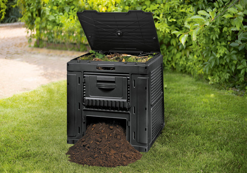 Czarny kompostownik z otwartą pokrywą, z którego wysypuje się ziemia. W tle widoczna jest zielona roślinność.