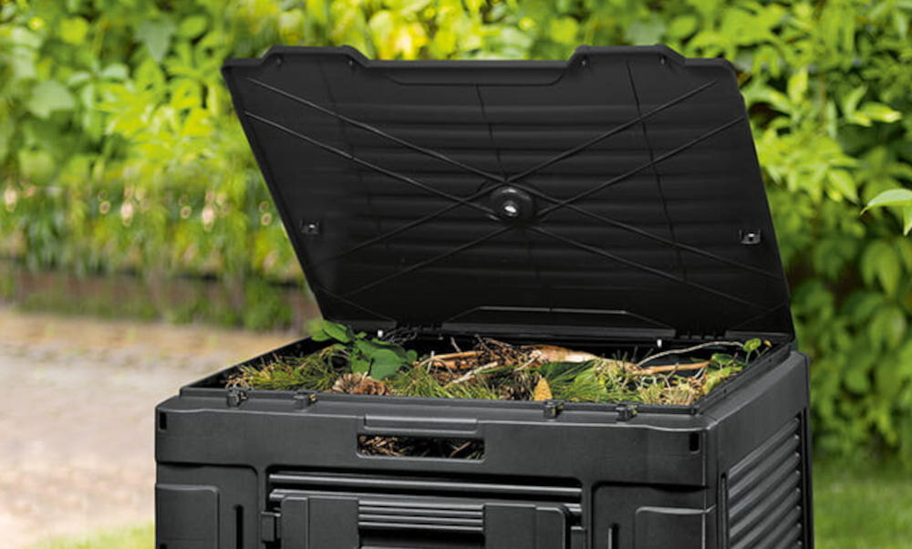Czarny kompostownik z otwartą pokrywą, wypełniony materiałem organicznym. W tle widoczne są zielone rośliny.