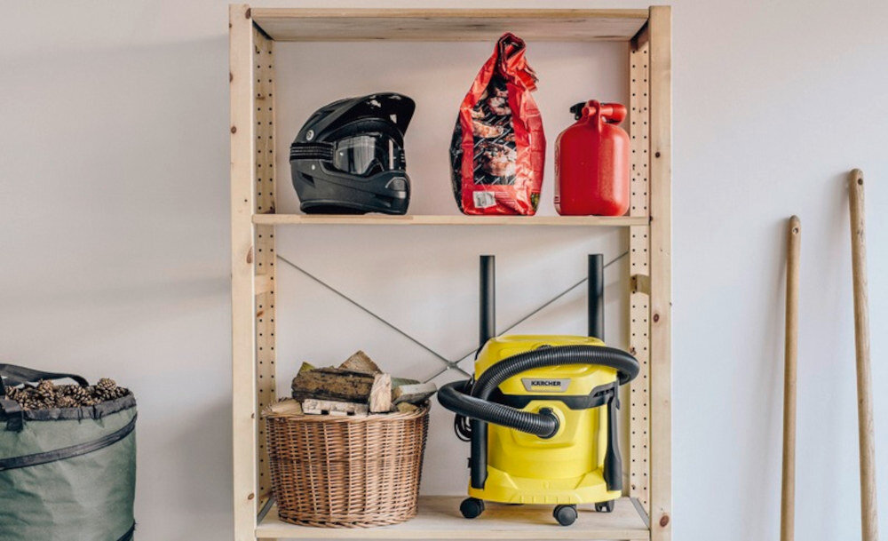 Na drewnianym regale znajdują się kask, worek z węglem drzewnym, czerwona kanisterka oraz żółty odkurzacz warsztatowy z logo KARCHER. Obok regału stoją drewniane kije, a na dolnej półce jest kosz z drewnem.