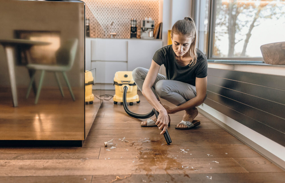 Osoba używa odkurzacza warsztatowego do sprzątania rozlanego płynu i resztek na drewnianej podłodze. W tle widoczny jest żółty odkurzacz oraz nowoczesna kuchnia.