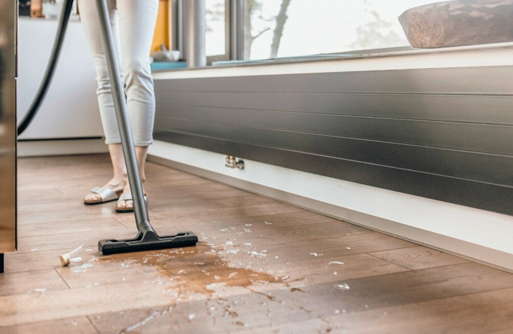 Osoba używa odkurzacza do sprzątania rozlanego płynu i rozbitego szkła na drewnianej podłodze. W tle widać okno i fragment ściany z listwą przypodłogową.