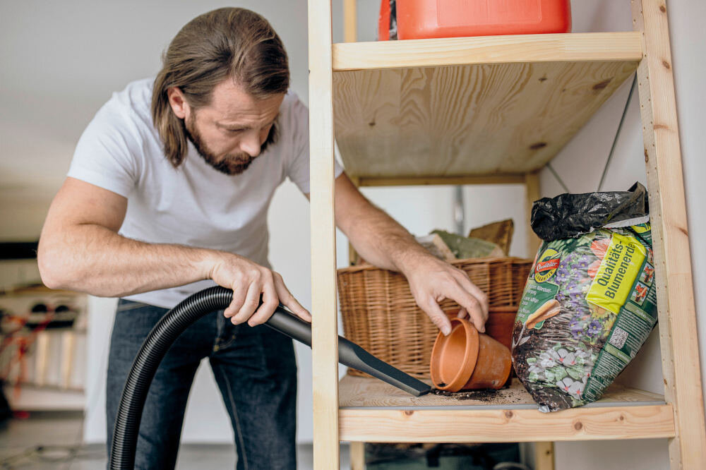 Osoba używa odkurzacza warsztatowego do sprzątania rozsypanej ziemi z półki. Obok znajduje się worek z ziemią ogrodniczą i przewrócona doniczka.