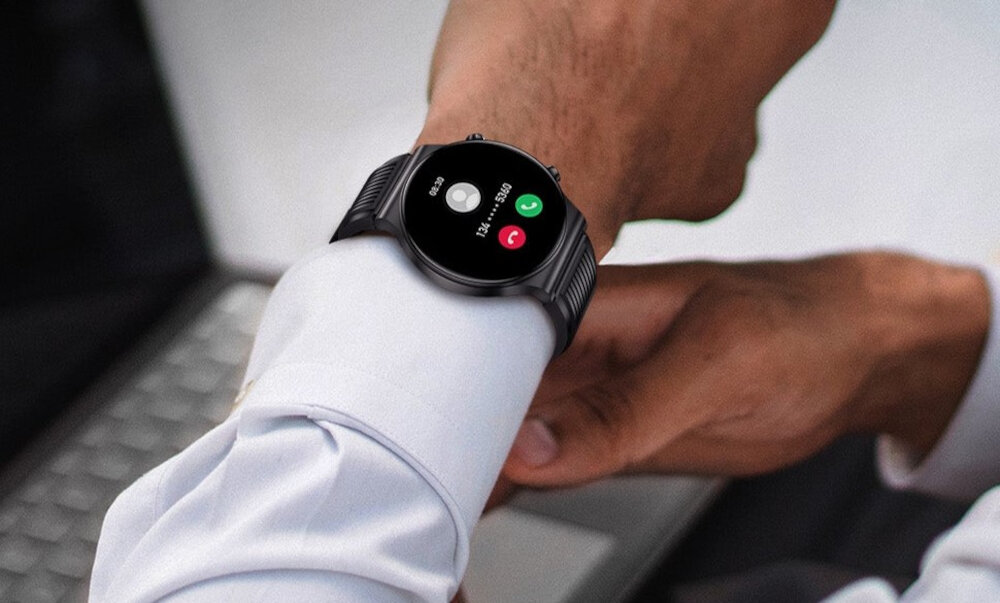 Osoba nosi czarny smartwatch z okrągłym wyświetlaczem, na którym widoczny jest ekran połączenia z przyciskami \'Odrzuć\' i \'Odbierz\'. W tle znajduje się klawiatura laptopa.